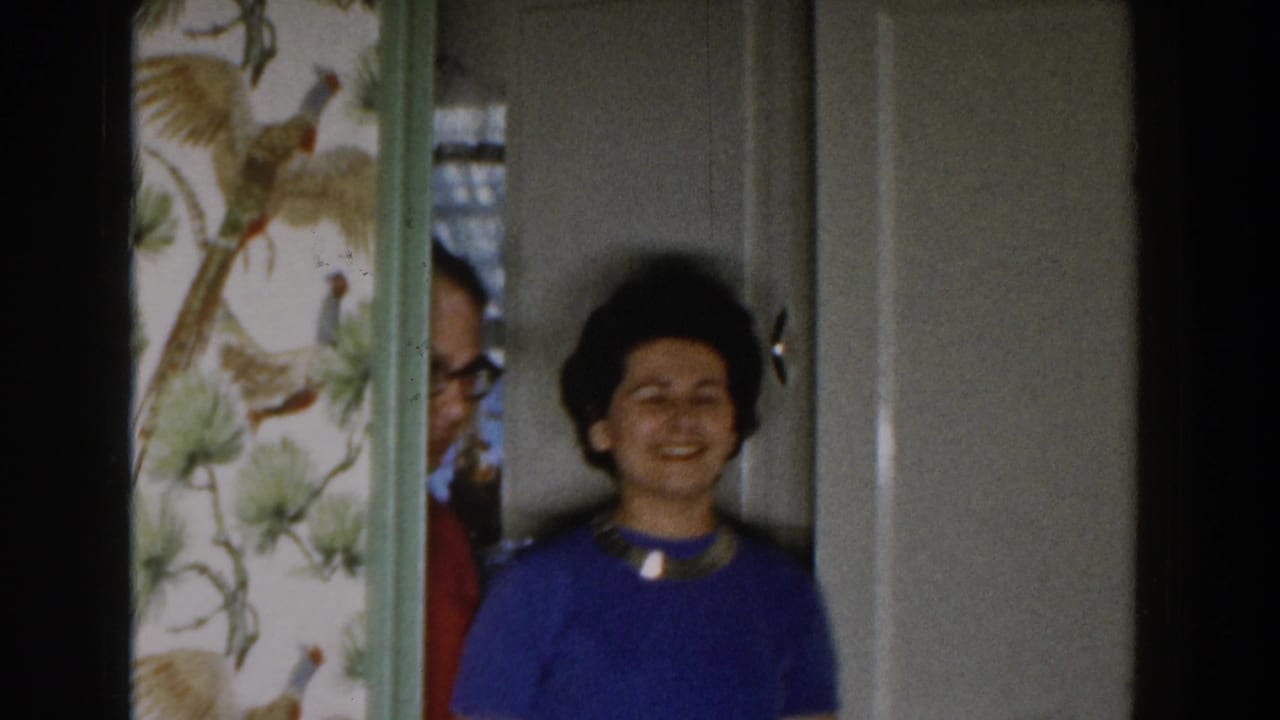 Woman in a blue top smiles near a doorway as a man partly appears behind her.