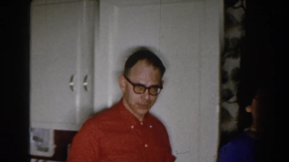 A man in glasses and a red patterned shirt stands near white cabinets inside a Michigan home, with another person partly visible at the edge.