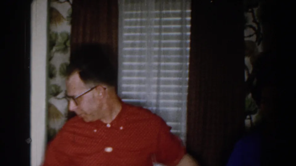 The man in the red patterned shirt turns to the side in a 1959 Michigan interior with venetian blinds, dark curtains, and bird-patterned wallpaper.