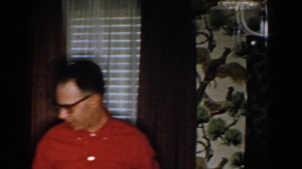 A man in glasses moves across a private Michigan home interior, with venetian blinds, dark curtains, patterned wallpaper, and a partial figure nearby.