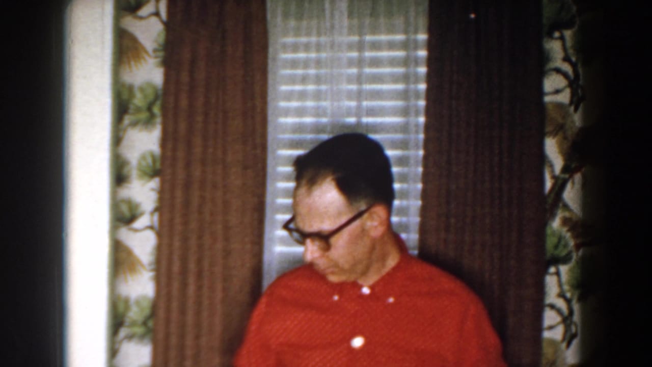 Man in red shirt and glasses stands by blinds and floral wallpaper.