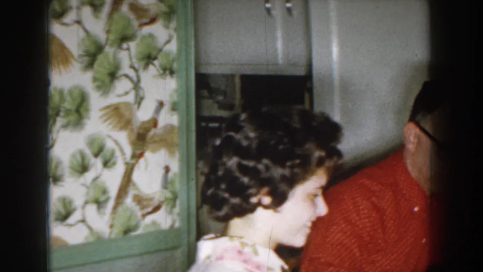 A woman with a curled 1950s hairstyle stands beside a man in red near white cabinets and floral wallpaper in the Michigan home.