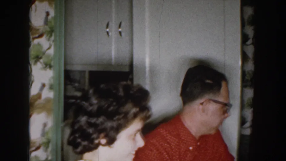 The woman appears in the foreground while the man in red turns near white kitchen cabinets and patterned wallpaper in the 1959 home movie.
