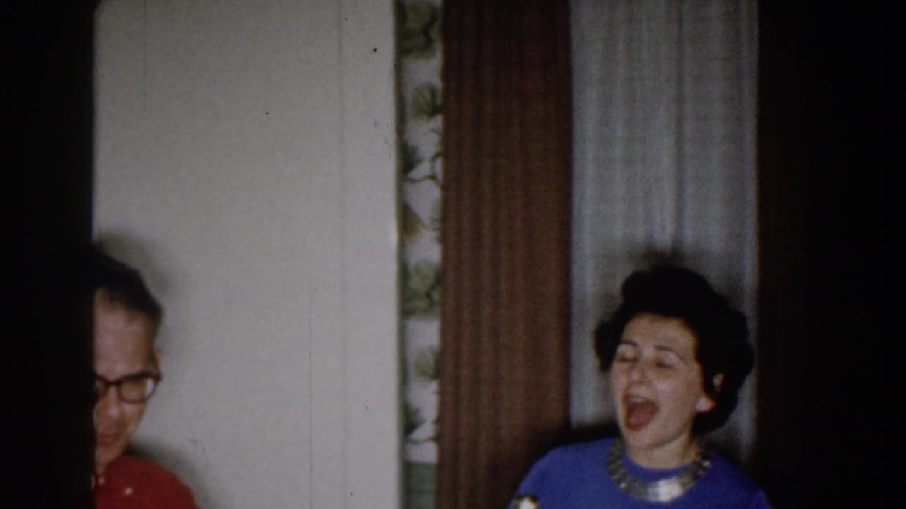 In Michigan 1959 home-movie footage, a woman in a blue sweater sings in a curtained room as a man is partly seen at frame left.
