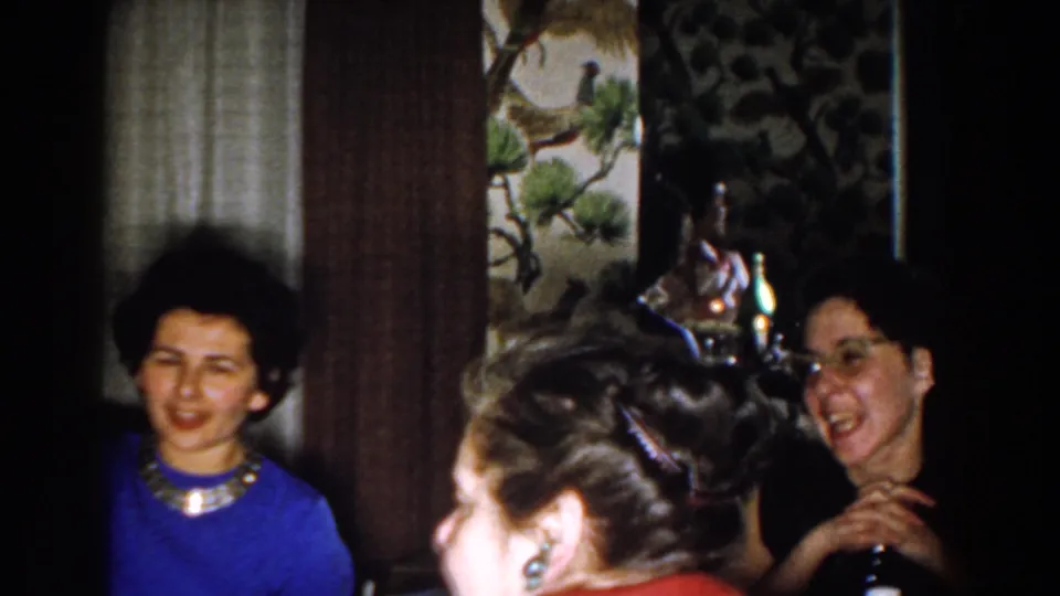 Michigan 1959 home-movie frame showing seated guests at a table, including a woman in blue and a woman with glasses in a dark top.