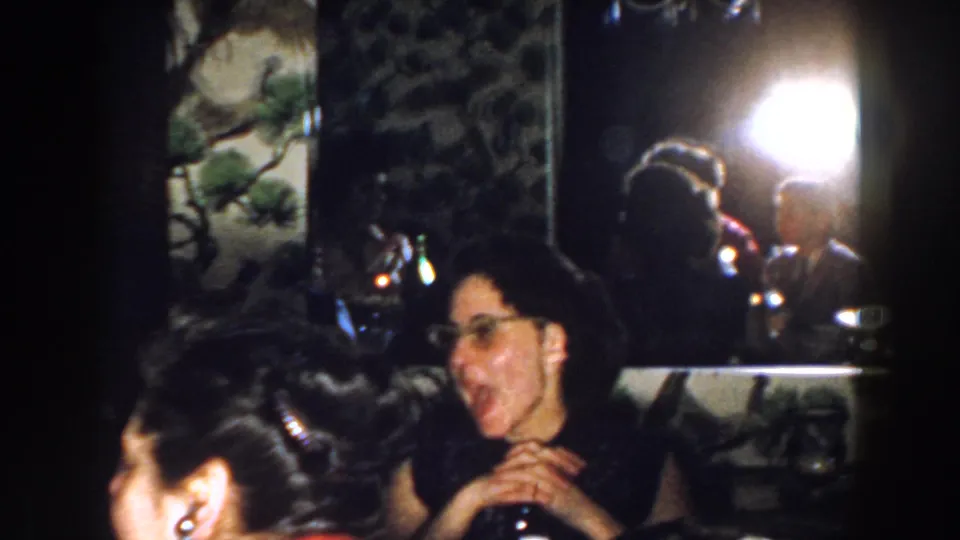 In the Michigan 1959 party scene, a woman with glasses sings near bottles and a mirror reflecting guests and a bright indoor light.
