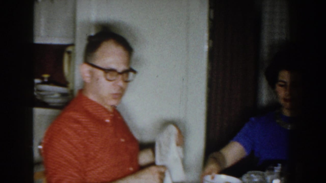 Man in a red patterned shirt and glasses sits at a dining table while a woman is partly visible nearby.