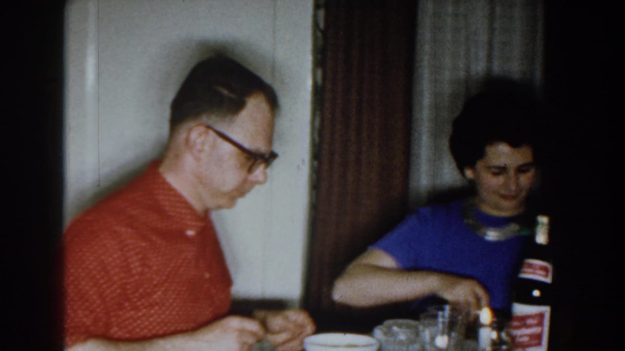 1959 Michigan home-movie frame showing two adults at a dining table with dishes, glasses, and a labeled bottle in view.