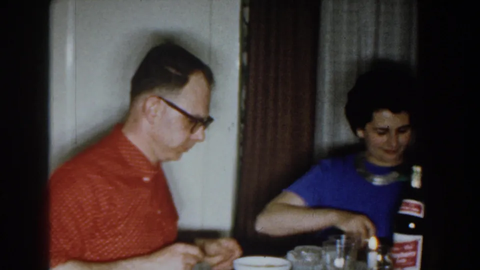 1959 Michigan home-movie frame showing two adults at a dining table with dishes, glasses, and a labeled bottle in view.