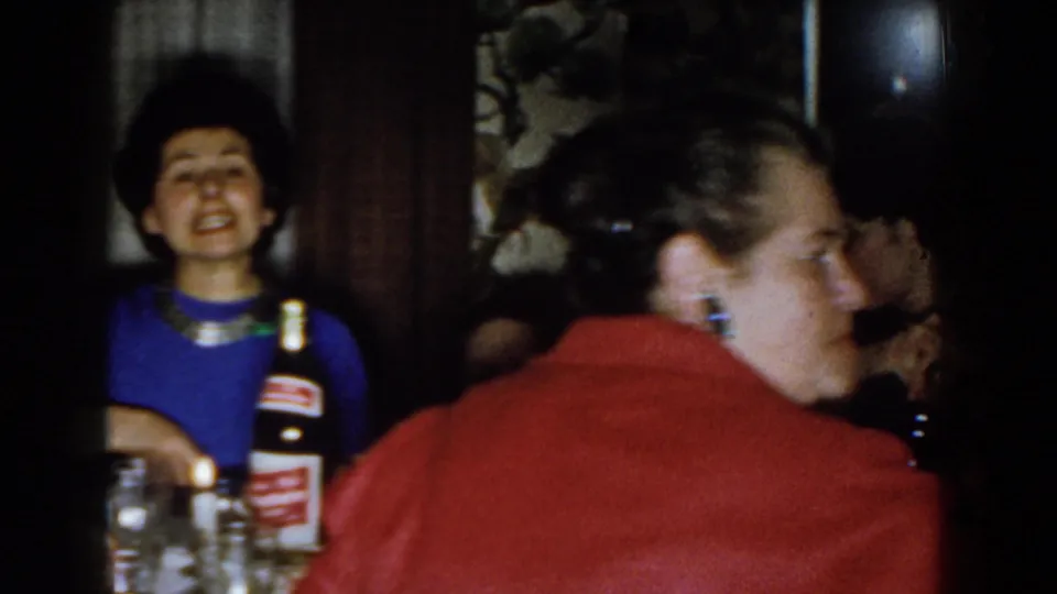 1959 Michigan 8mm frame of a woman at a home dining table, with a labeled bottle and another adult close to the camera.
