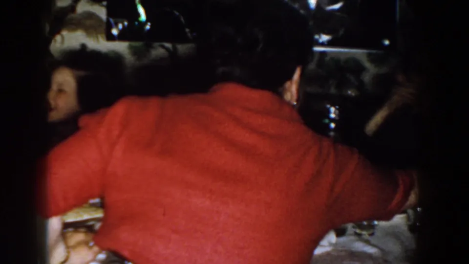1959 Michigan home interior frame showing the back of a red-shirted adult at a crowded dinner table with other faces partly visible.