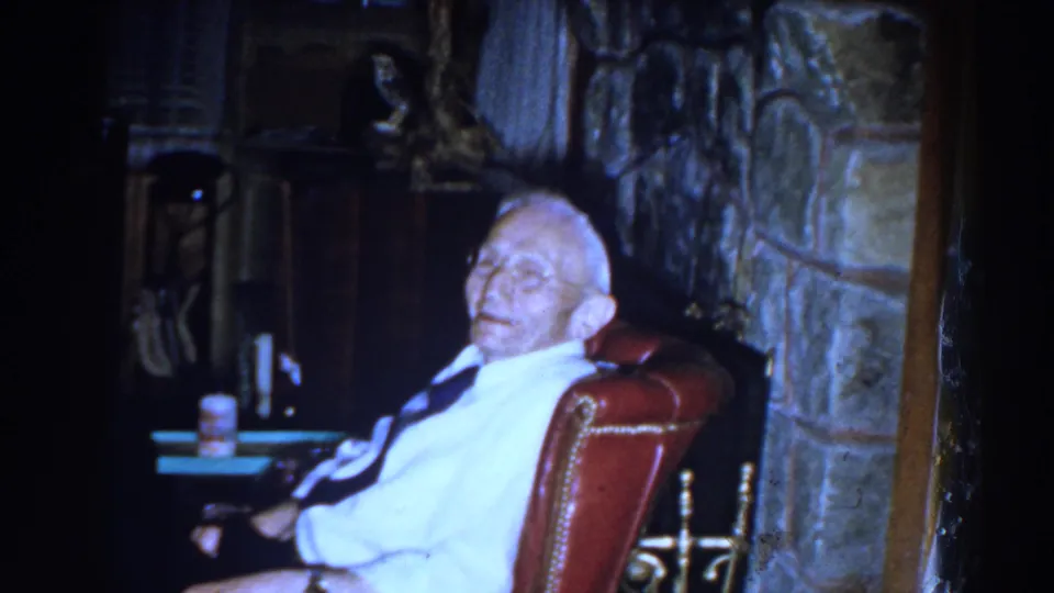 In a 1959 Michigan home interior, an elderly man in a white shirt and dark tie sits in a red leather chair near a stone fireplace.
