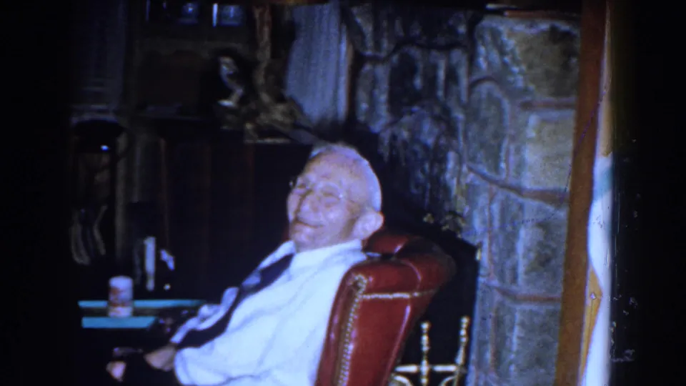 The seated elderly man appears to smile toward the camera in a dim 1959 Michigan interior with a red chair and stone fireplace.