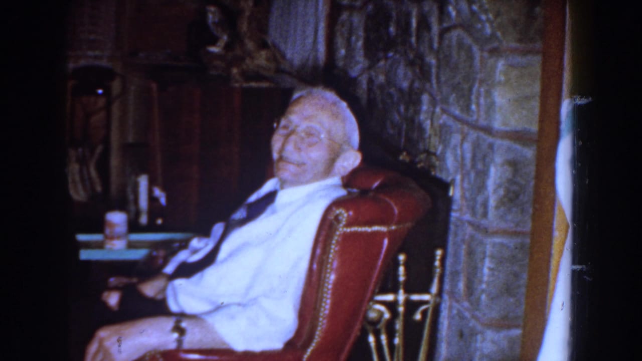 Elderly man in a white shirt and tie sits in a red leather chair near a stone fireplace.