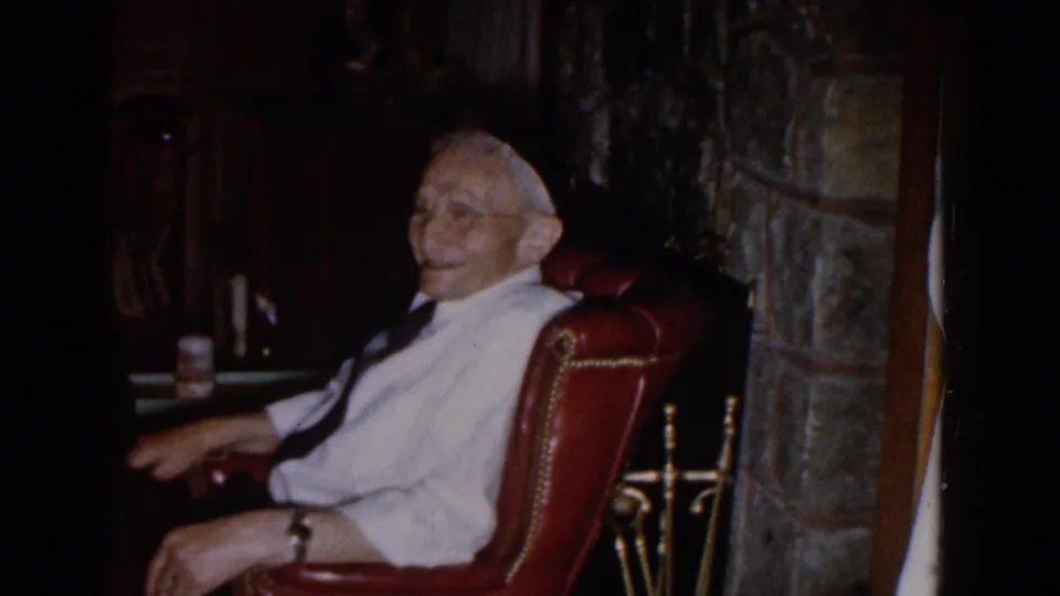 The elderly man smiles toward the camera while seated in a red leather chair in a dim 1959 Michigan home interior.
