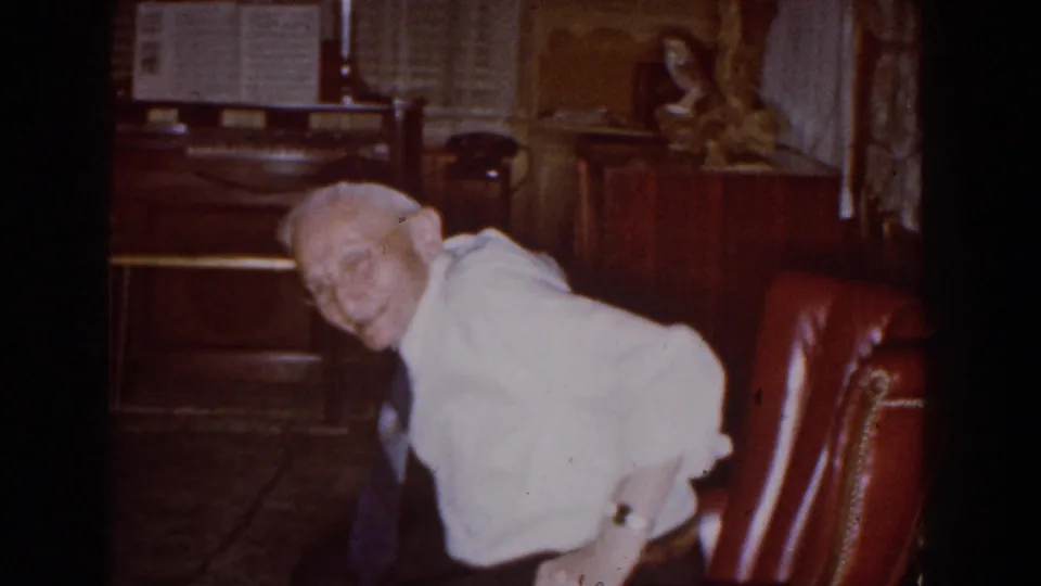 At the end of the clip, the man leans forward from the red armchair in a paneled Michigan home interior.