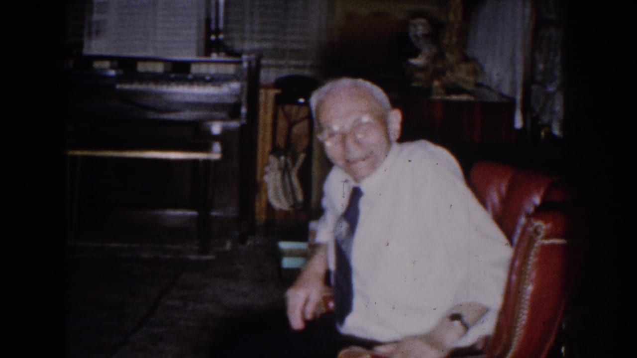 In a 1959 Michigan home interior, an elderly man smiles from a red chair near a piano, blinds, and dark furnishings.
