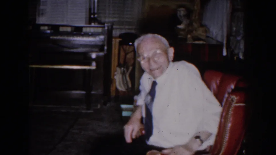 In a 1959 Michigan home interior, an elderly man smiles from a red chair near a piano, blinds, and dark furnishings.