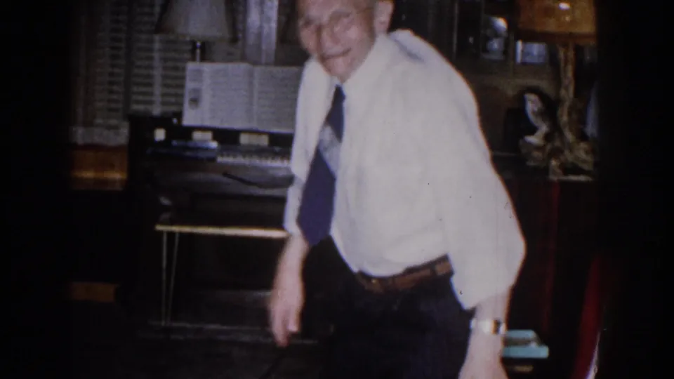 The elderly man stands beside the red chair, smiling toward the camera in the 1959 Michigan home setting.
