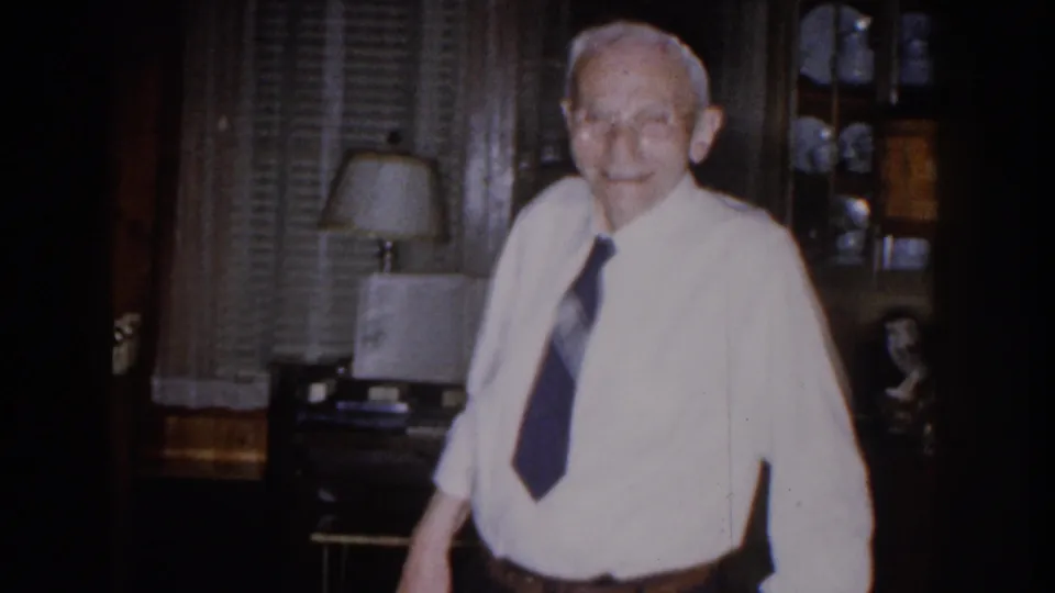 The man stands closer in the dim home interior, with blinds, a lamp, shelves, and wood furnishings behind him.