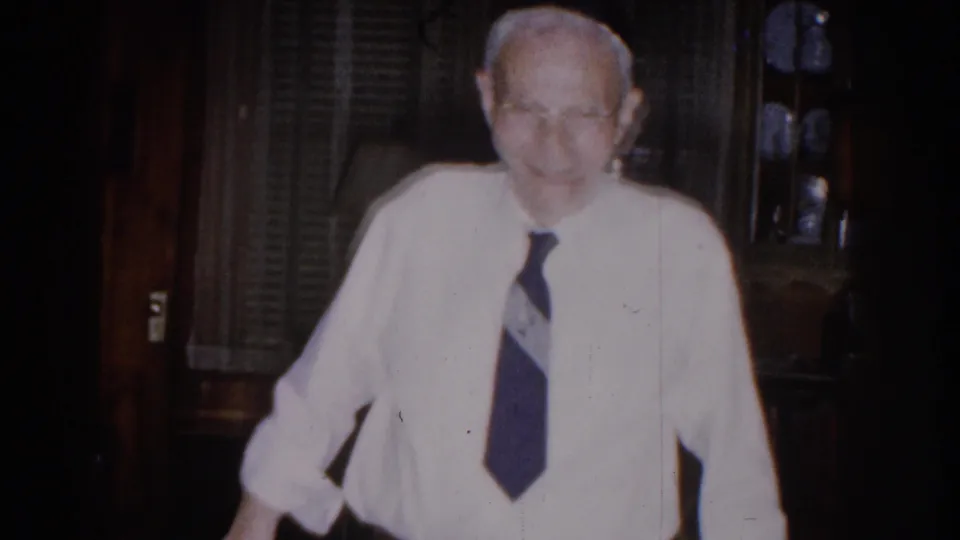 The elderly man faces the camera in white shirt and tie, framed by blinds, shelves, and dark wood details.