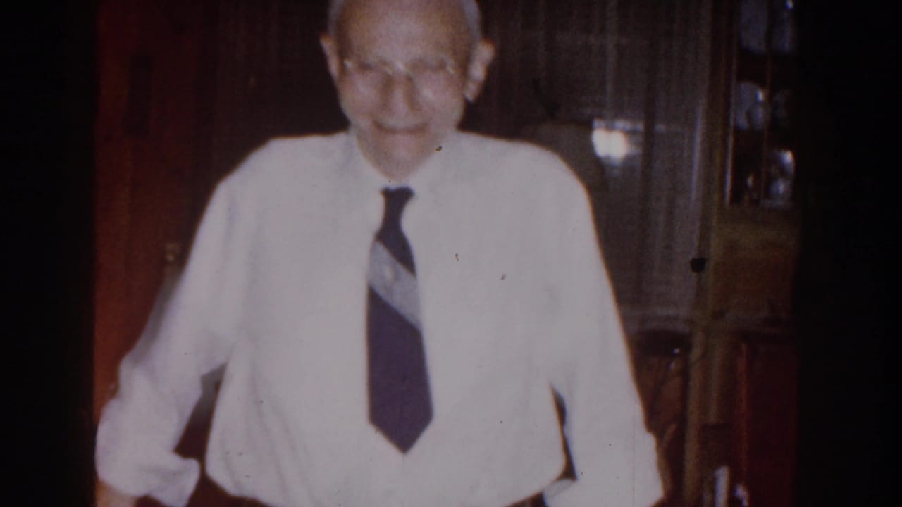 Elderly man in a white shirt and dark tie smiling in a dim living room.