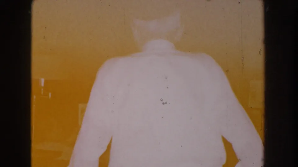 The elderly man is seen from behind in a white shirt as the warm, overexposed 8mm frame obscures much of the room detail.