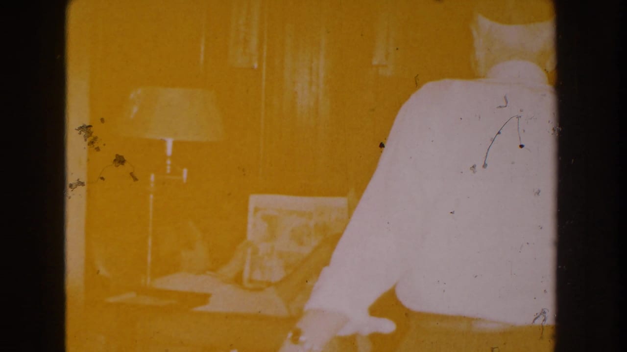 1959 Michigan home-movie frame with strong yellow exposure showing a living room, seated woman, newspaper, lamp, and man moving past camera.