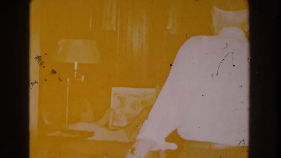 1959 Michigan home-movie frame with strong yellow exposure showing a living room, seated woman, newspaper, lamp, and man moving past camera.