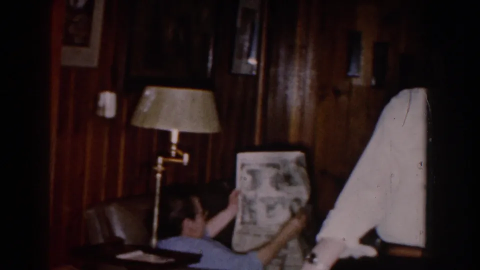 1959 Michigan living room scene with a woman reading a newspaper on a couch while a man in a white shirt and dark hat stands beside her.