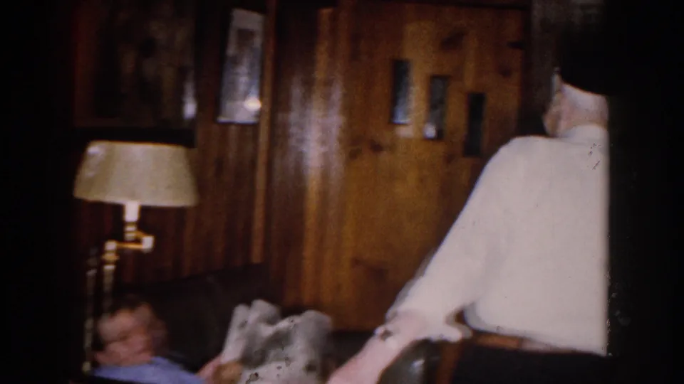 1959 Michigan home interior with a man in a white shirt and dark hat walking away from the camera beside a couch and lamp.