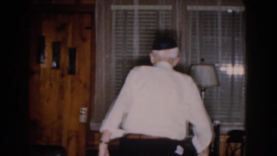 1959 Michigan home-movie frame showing a man from behind near window blinds in a wood-paneled living room with a lamp at right.