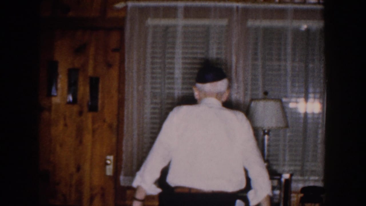 In a 1959 Michigan home-movie frame, an elderly man in a white shirt and dark head covering walks away from the camera in a dim living room.