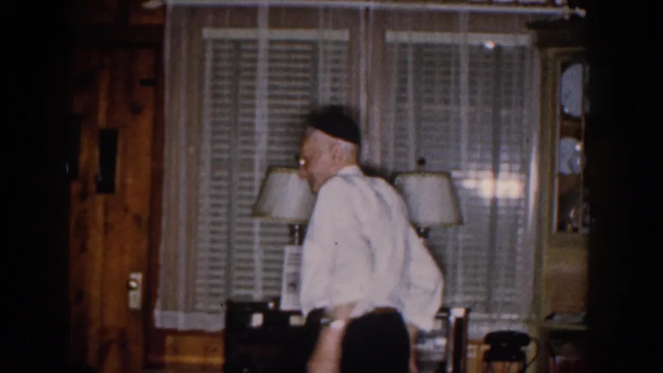 The elderly man appears in profile in the Michigan living room, wearing a white shirt and dark head covering beside curtained windows and lamps.