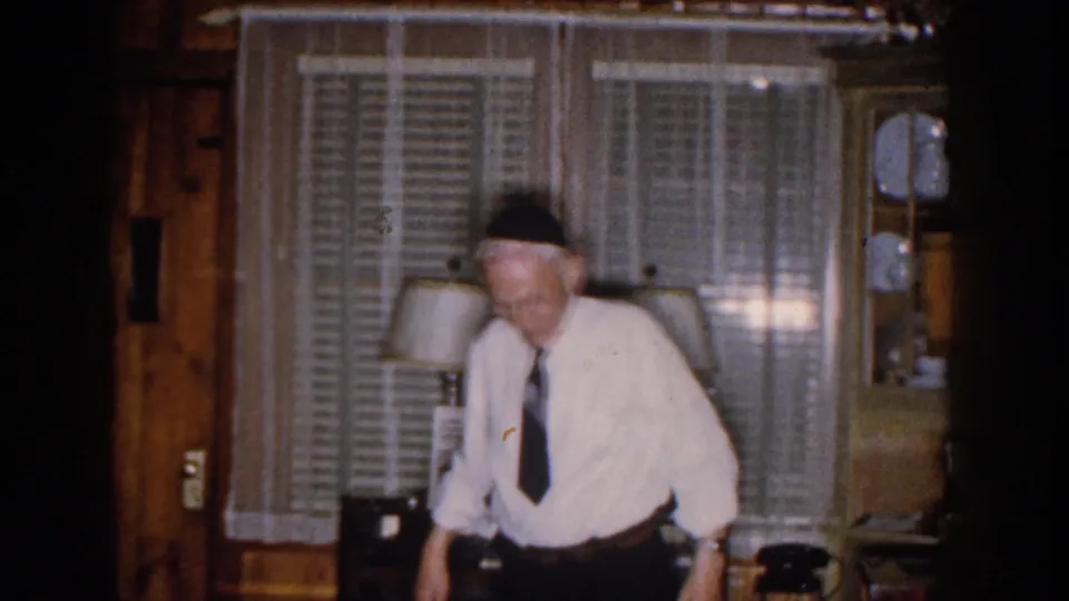 The man lowers his gaze in the dim 1959 Michigan interior, with his dark tie visible against a white shirt near lamps and curtains.