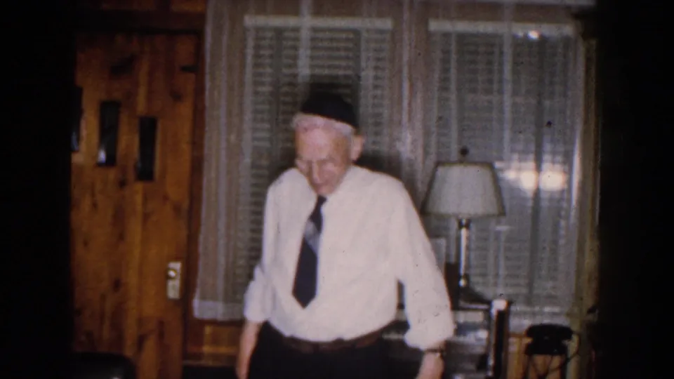 The elderly man faces the camera in a Michigan home interior, wearing glasses, a white shirt, dark tie, and dark head covering.