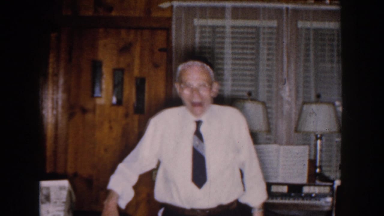 Older man in white shirt and dark tie standing in a wood-paneled room.