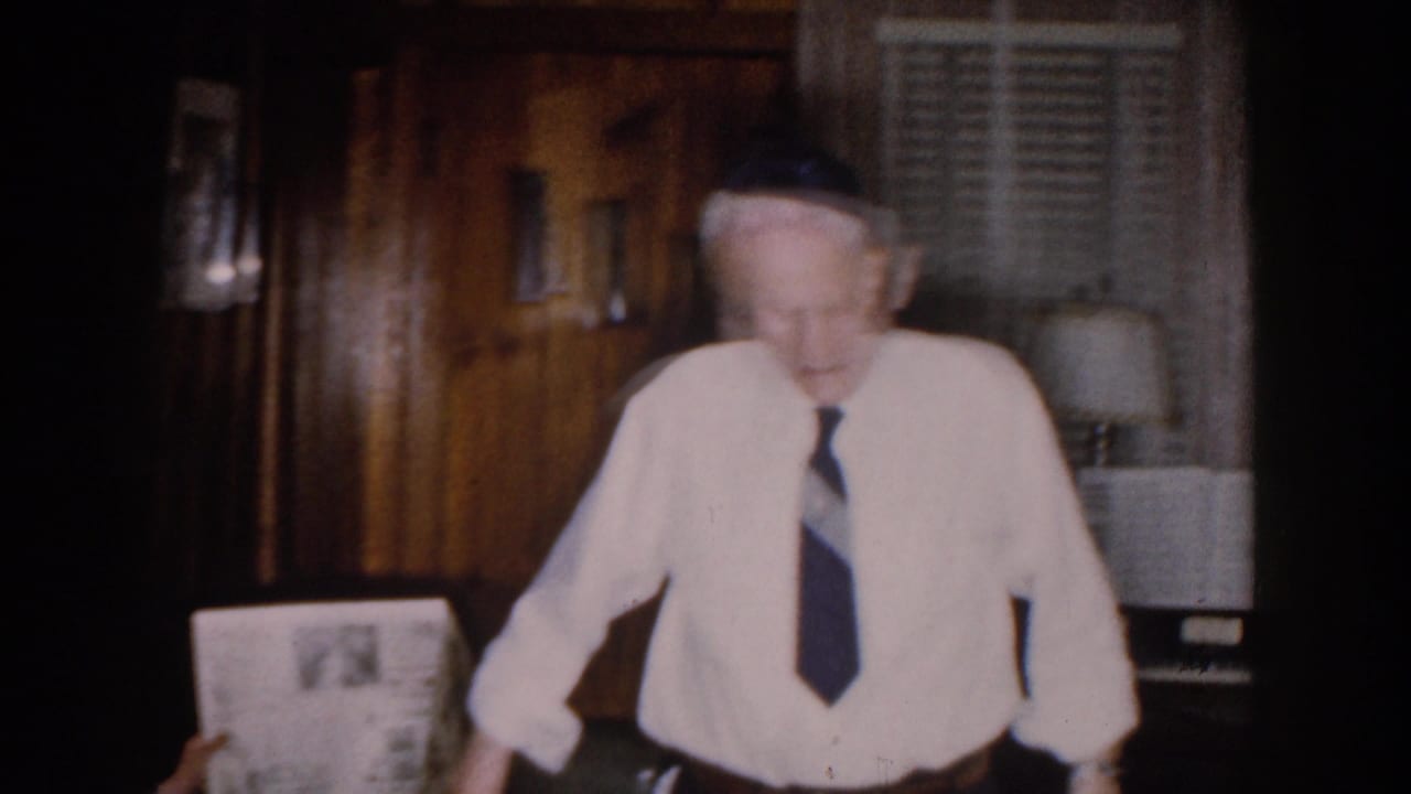 Older man leaning forward in a wood-paneled interior with lamps behind him.