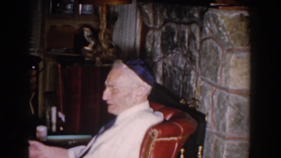 Older Jewish man in a yarmulke seated beside a stone fireplace in a private Michigan home interior, 1959.