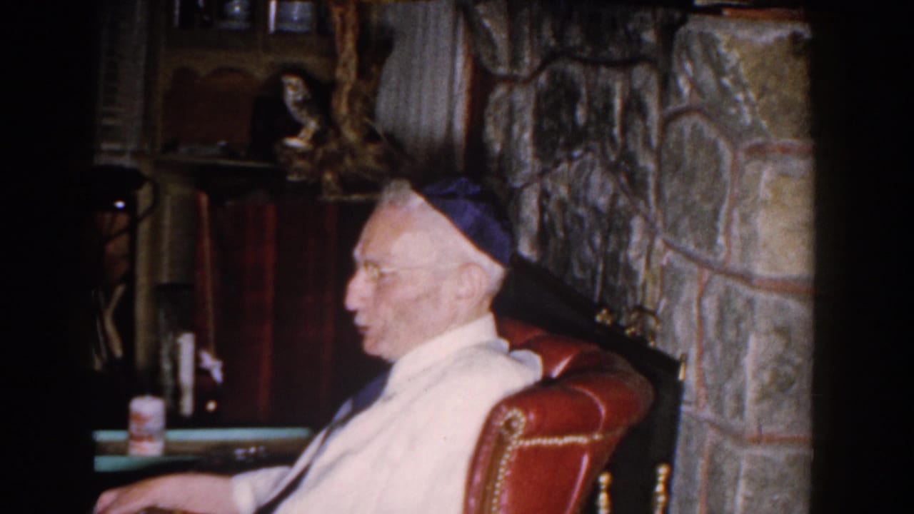 In a 1959 Michigan home interior, an elderly man in a white robe and blue skullcap sits in profile near a stone fireplace.