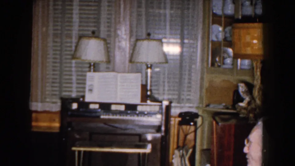 A wider living room view shows a piano with sheet music, table lamps, shutters, shelves, and the seated man's head at the lower edge.