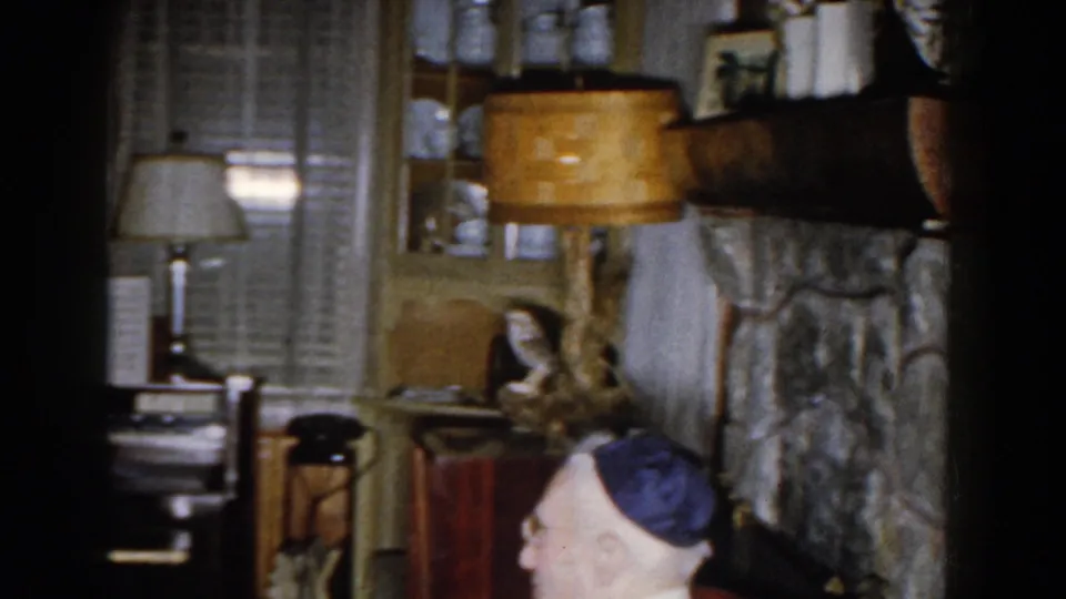 The 1959 Michigan interior frames the elderly man low in the shot, with stone fireplace, piano, lamp, shelves, and decor around him.