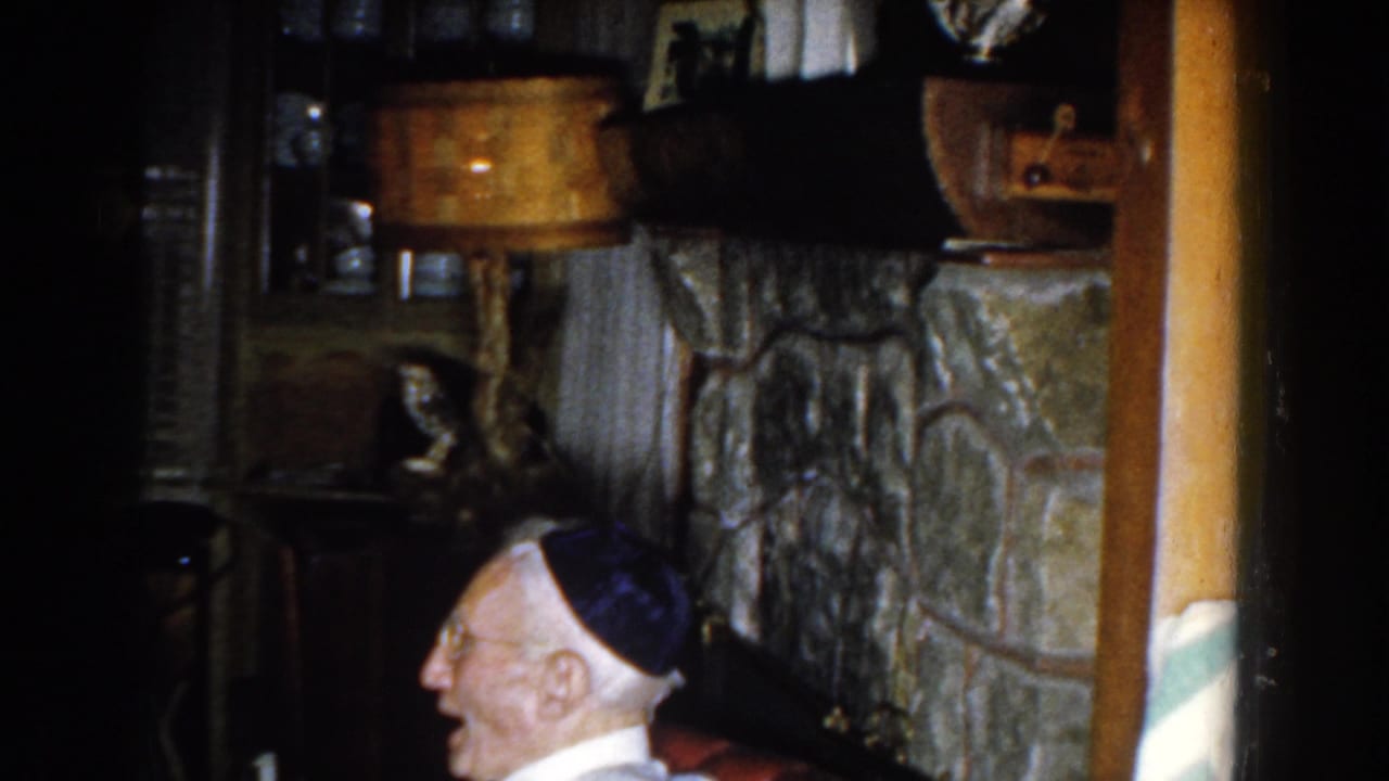 Elderly man in dark cap seated near a stone fireplace in a dim living room.