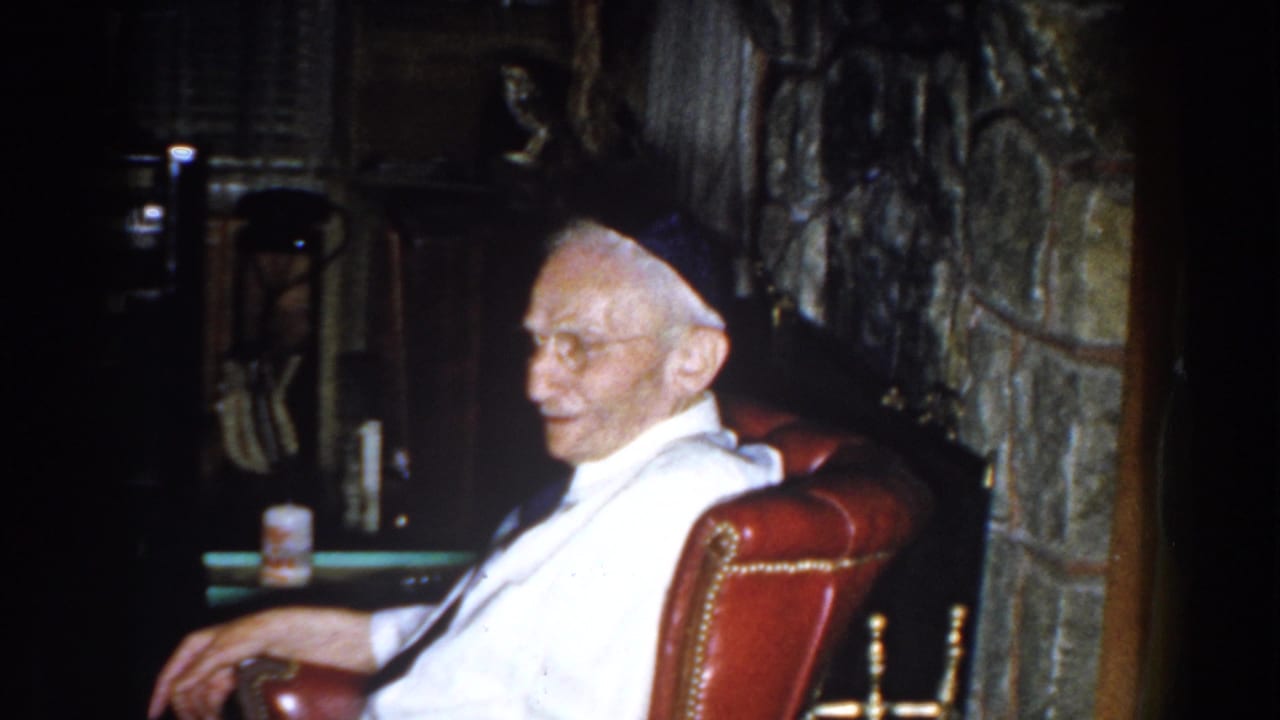 Elderly man in white shirt sitting in a red armchair beside a stone fireplace.