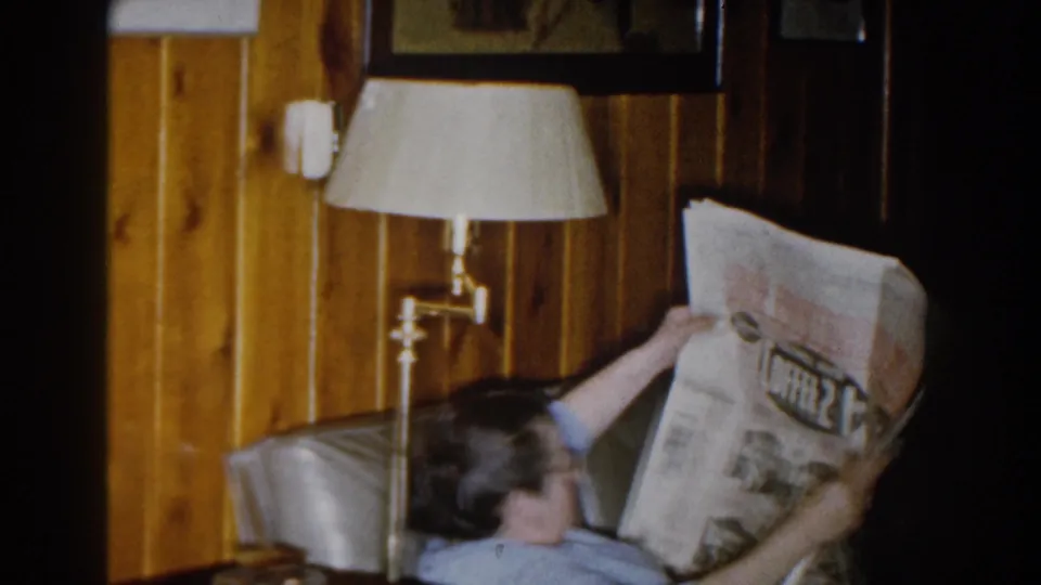 A person reads a newspaper on a sofa in a wood-paneled Michigan living room from the 1959 home movie.