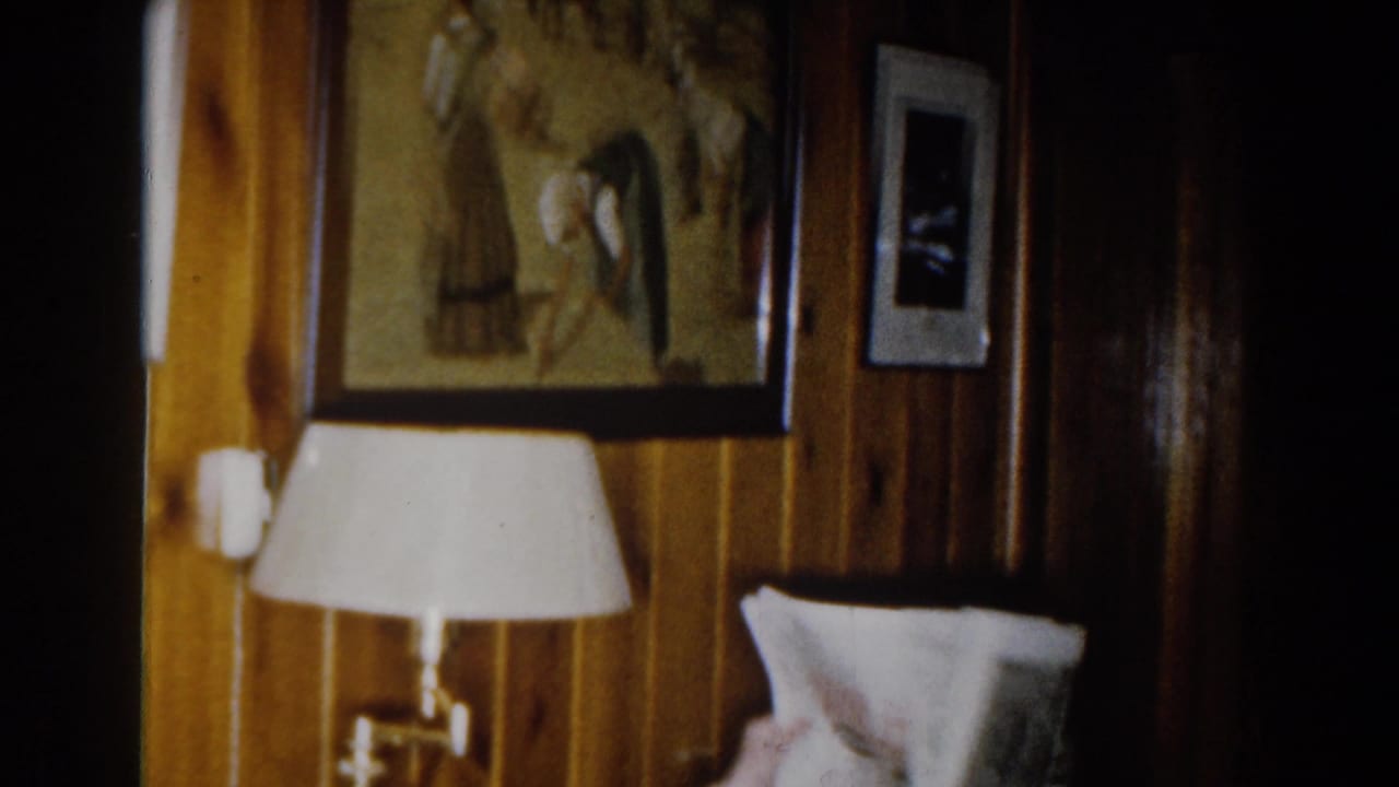 1959 Michigan home-movie frame showing a wood-paneled interior with framed art, a small wall photo, a white-shaded lamp, and part of a sofa.