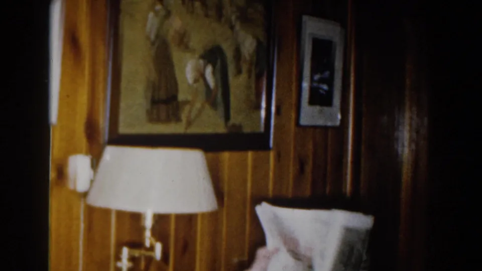 1959 Michigan home-movie frame showing a wood-paneled interior with framed art, a small wall photo, a white-shaded lamp, and part of a sofa.