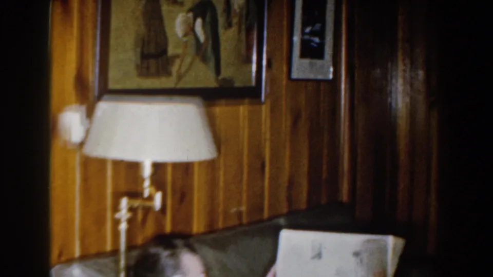 1959 Michigan home-movie frame with a person partly visible on a dark sofa below framed wall art and a white-shaded lamp in a paneled room.