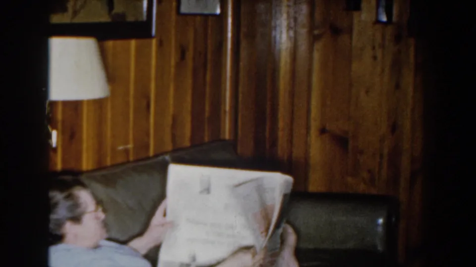 1959 Michigan home-movie frame showing a seated person in a light blue shirt reading a newspaper on a dark sofa in a wood-paneled room.