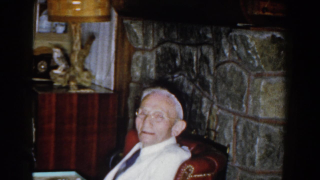 Elderly man with glasses seated in a red armchair near a stone fireplace.